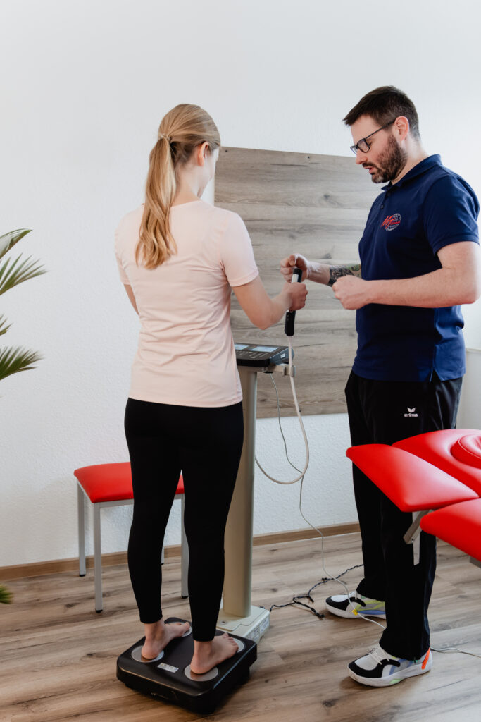 Patient auf Körperanalysewaage. Mitarbeiter erklärt Messung.